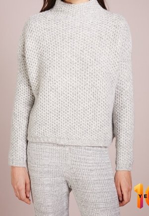 Femme portant un pull en tricot texturé gris clair et un pantalon en tricot côtelé assorti, debout devant un fond rose pâle.