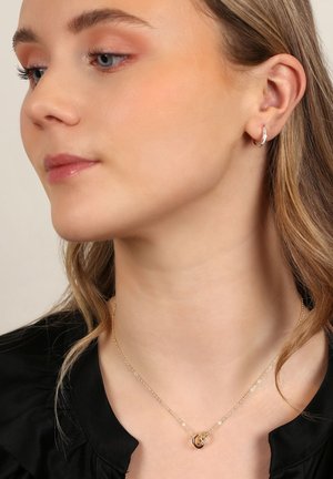 Guldkedja med en cirkulär berlock, bärs mot bar hud, tillsammans med små silverhänge. En svart volangblus är delvis synlig.