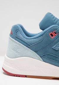 Talons de baskets en daim bleu et gris clair avec des accents rouges, semelle intermédiaire blanche estampillée "ENERP" et semelle extérieure en caoutchouc texturé rouge et marron.