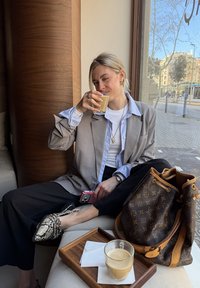Femme en blazer gris et pantalon noir assise près de la fenêtre, faisant un clin d'œil en tenant un verre de café, avec un sac de créateur et un autre café sur un plateau.