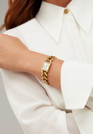 Montre-bracelet de couleur dorée avec un cadran rectangulaire et un bracelet à maillons, portée sur un bras vêtu d'une chemise en tissu blanc léger et fluide.