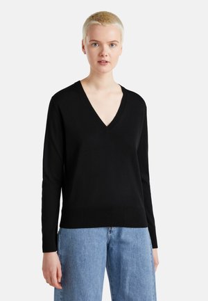 Vrouw met kort blond haar, gekleed in een zwarte V-halstrui en lichtblauwe spijkerbroek, tegen een effen witte achtergrond.