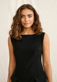 Junge Frau mit lockigem braunem Haar, die ein ärmelloses schwarzes Oberteil trägt, vor einem einfarbigen hellen Hintergrund steht und in die Kamera blickt.