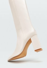 Kniehohe, weiße Lederstiefel mit spitzer Zehenpartie und einem dicken beigen Absatz. Glatte Textur mit dezenten Nähten und Logo auf der Sohle.