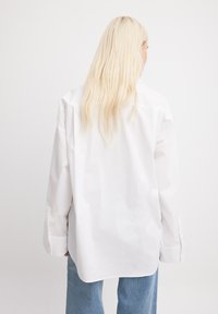 Gari plāniem piedurknēm baltā kokvilnas krekls ar brīvu piegriezumu, atpakaļ yoke detaļu un pogu aprocēm, apvienots ar gaiši ziliem džinsiem.