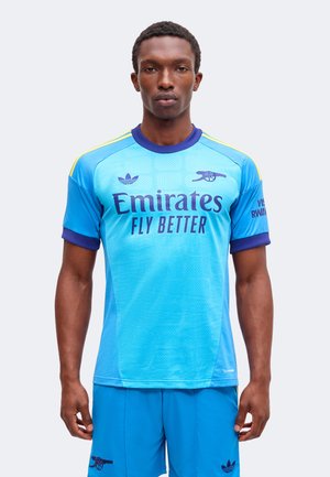 Mężczyzna ubrany w niebieski strój piłkarski Arsenalu z logo "Emirates Fly Better", stojący na jednolitym jasnym tle.