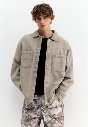 Ung mand med krøllet hår iført en beige jakke med knapper over en sort skjorte og camouflagemønstret bukser, stående med hænderne i lommerne.