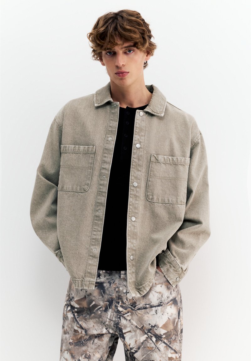 Mlad moški z kodrastimi lasmi, oblečen v bež jopo z gumbi prek črne majice in hlače s kamuflažnim vzorcem, stoji z rokami v žepih.