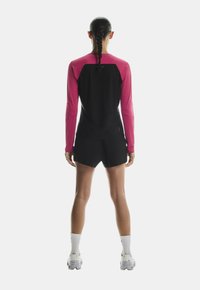 Dugih rukava sportska majica u ružičastoj i crnoj boji s uskim krojem. Uparen s crnim kratkim hlačama i bijelim čarapama. Pogled s leđa, glatka tekstura.