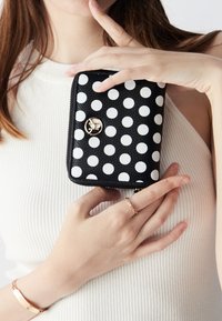 Portafoglio nero con pois bianchi, superficie texturizzata, accentuazione del logo dorato, chiusura con zip, tenuto in mano con una carnagione chiara su uno sfondo bianco.