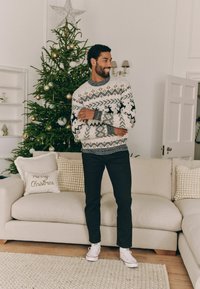 Gebreide trui in crème met zwarte en gouden patronen, donkere broek en witte sneakers. Gezellige woonkamer met een kerstboom.