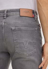 Jeans in denim grigio con etichetta in pelle che riporta la scritta "JOOP! JEANS." Presentano un design distintivo della tasca posteriore con un motivo a cucitura zigzag.
