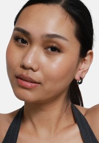 Schwarze Creolenohrringe mit strukturierten Details, getragen von einem Model mit glatter Haut, das ein natürliches Make-up und dunkles Haar aufweist.