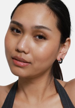 Boucles d'oreilles créoles noires avec des détails texturés, portées par un modèle avec un teint lisse, arborant un maquillage naturel et des cheveux foncés.