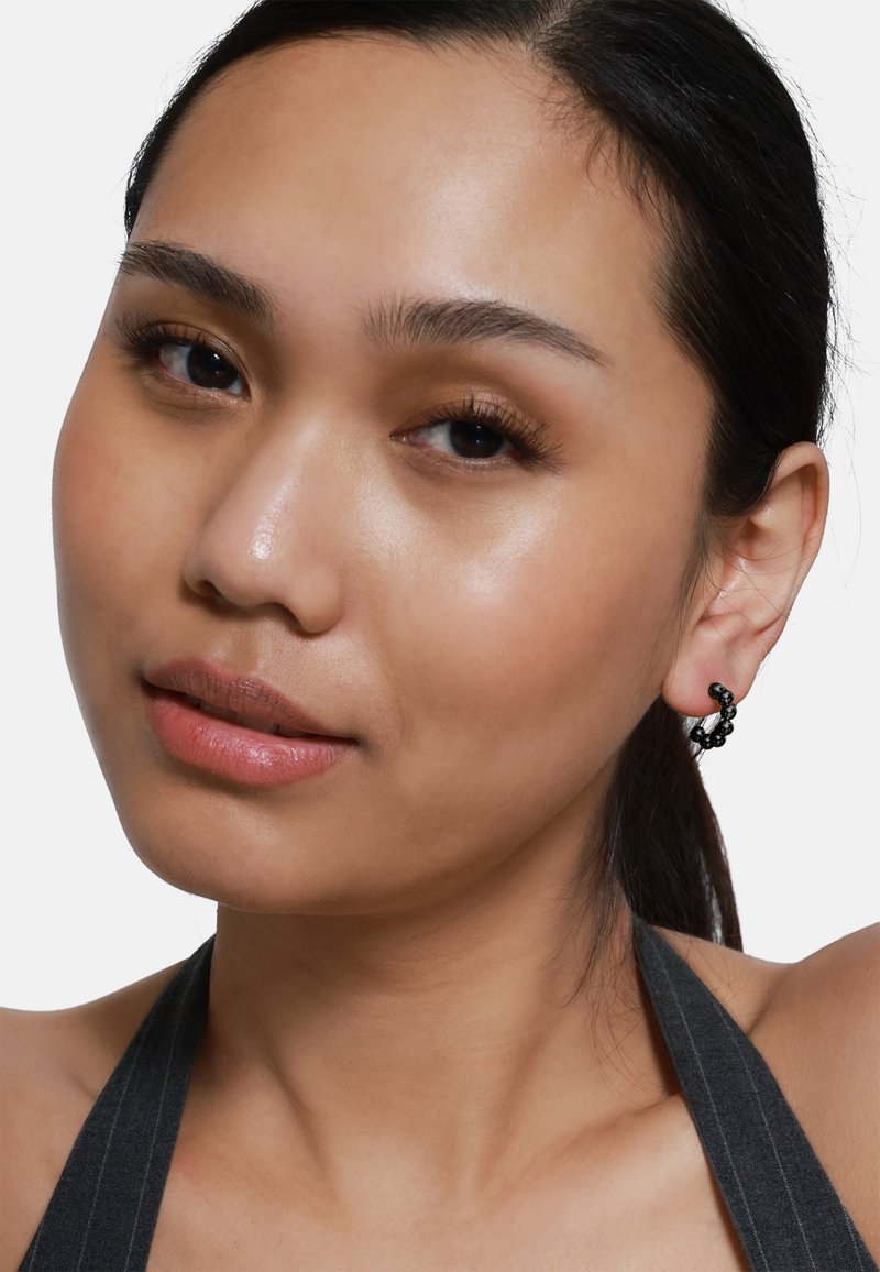 Schwarze Creolenohrringe mit strukturierten Details, getragen von einem Model mit glatter Haut, das ein natürliches Make-up und dunkles Haar aufweist.