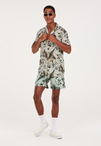 Camicetta a maniche corte con bottoni e pantaloni abbinati in verde con motivi di foglie tropicali. Gli accessori includono occhiali da sole e calzini bianchi con scarpe slip-on.