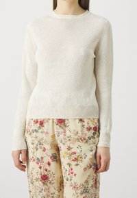 Pull en maille beige clair à manches longues, col rond et poignets côtelés, associé à un pantalon à motifs floraux sur un fond neutre.