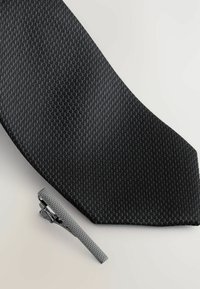 Crni kravata s teksturom uskog oblika pored srebrne kopče za kravatu koja ima teksturiranu površinu i elegantan dizajn.