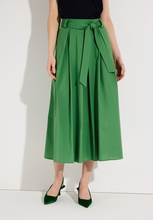 Femme portant une jupe midi verte plissée avec une ceinture nouée à la taille, assortie à des escarpins slingback pointus vert foncé.
