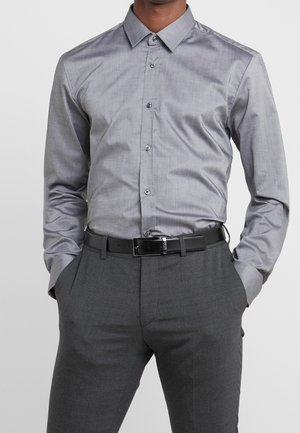 Mann trägt ein eng anliegendes graues Hemd mit Knöpfen, das in dunkelgraue Hose mit schwarzem Gürtel gesteckt ist, Hände in den Taschen, vor einem schlichten Hintergrund.