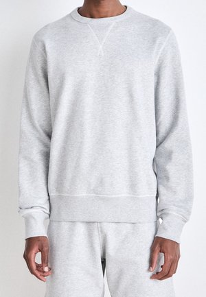 Mand iført lys grå langærmet rundhalset sweatshirt og matchende joggingbukser, stående foran en ensfarvet hvid baggrund.