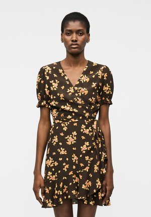 Mujer vistiendo un vestido cruzado floral de color marrón oscuro con mangas cortas abullonadas y dobladillo con volantes sobre un fondo claro y liso.