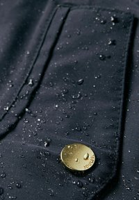 Jūras zila ūdensnecaurlaid�īga auduma virsma ar ūdens pilēm, greznu zelta pogu ar iegravētu tekstu "JOULES". Tekstūrēta virsma ar redzamām šuvju detaļām.