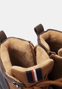 Botas de invierno marrones con forro de felpa beige, cremallera lateral y tiradores de tela, con un detalle de rayas azules, rojas y blancas en la lengua.
