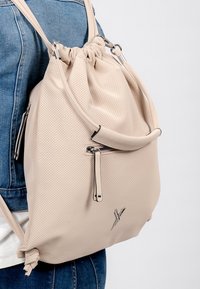 Mochila perforada beige con un cierre de cordón, correas ajustables y un detalle de cremallera plateada en el frente. Material texturizado.