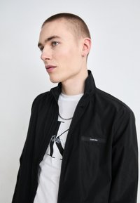 Mlad moški s kratko odrezanimi lasmi, oblečen v črno jakno Calvin Klein čez belo majico Calvin Klein, obrnjena levo proti beli ozadju.