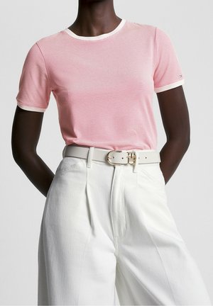 Kobieta ubrana w różowo-białą koszulę z cienkimi paskami i krótkim rękawem, włożoną w białe spodnie z wysokim stanem, przewiązane białym paskiem ze złotą klamrą.