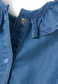 Prenda de mezclilla con un cuello volado; presenta un botón a presión plateado y un cierre de botón de color azul claro. La textura de la tela es suave y ligeramente desgastada.