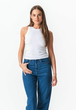 Nő hosszú barna hajjal, fehér ujjatlan felsőt és kék, magas derekú farmert viselve, fehér háttér előtt áll.