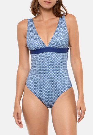 Femme portant un maillot de bain une pièce bleu et blanc à motifs, avec un profond décolleté en V et une bande unie bleu marine sous la poitrine, debout devant un fond uni.