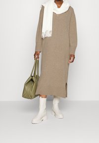 Beige stickad klänning med V-ringning, långa ärmar, sidoflik. Kombinerad med en vit scarf och en grön läderväska. Vita kängor med tjock sula och ankellängd.