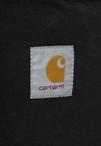 Etiqueta de tela blanca con el logo "C" en naranja y "carhartt" en texto marrón; unida a un fondo negro, con bordes cosidos.
