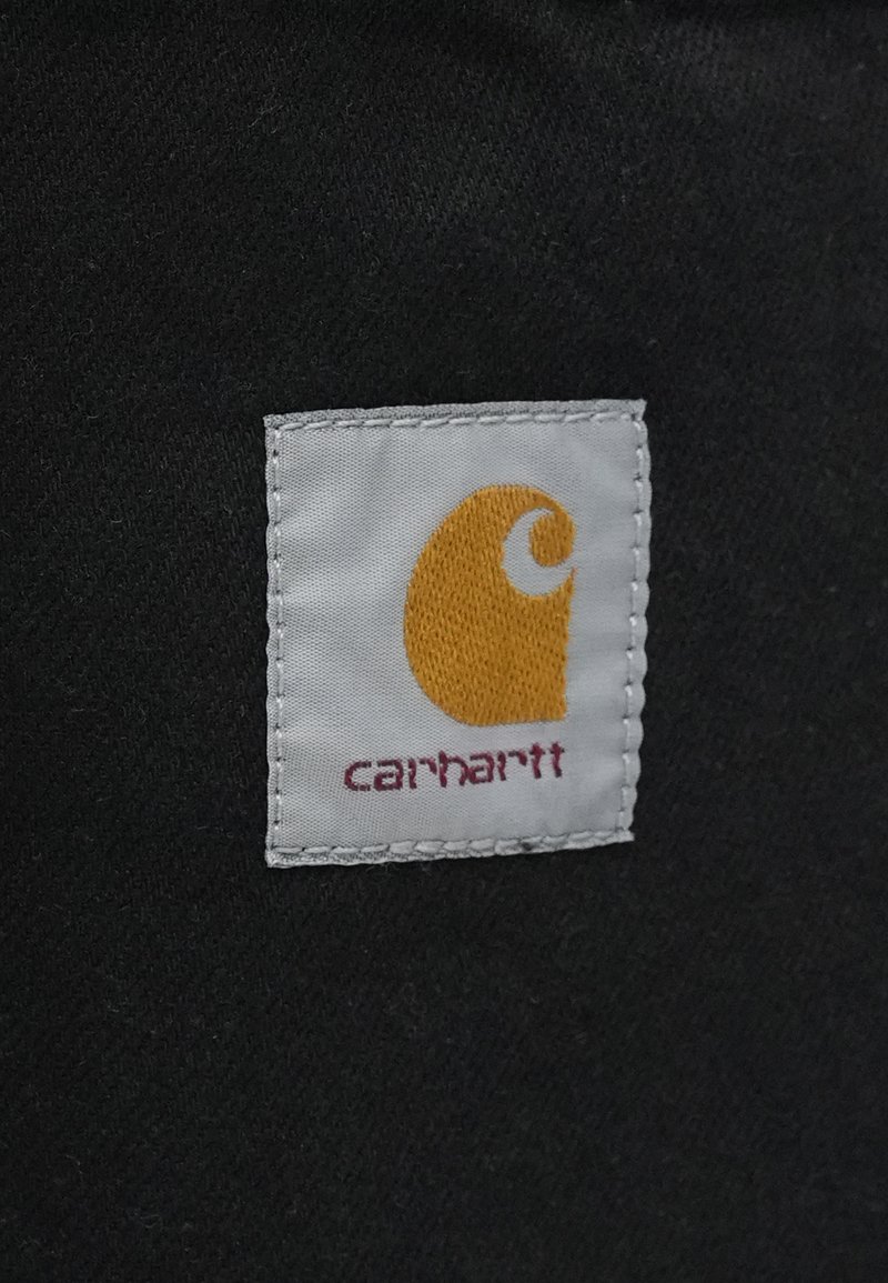 Weißes Stoffetikett mit orangefarbenem "C"-Logo und "carhartt" in kastanienbraunen Buchstaben; angebracht auf einem schwarzen Hintergrund mit genähten Kanten.