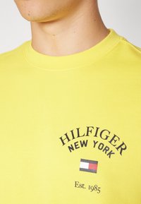 Gelbes Baumwoll-T-Shirt mit kurzen Ärmeln. Mit schwarzem Text "HILFIGER NEW YORK" und einem kleinen Logo-Graphic in Rot, Weiß und Blau. Rundhalsausschnitt.