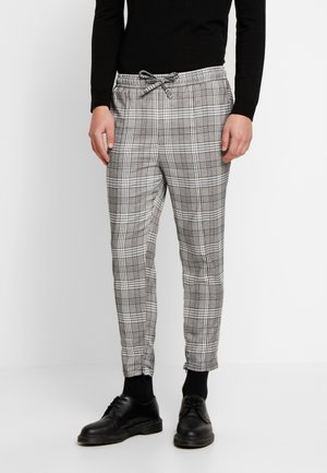 Mann trägt graue und weiße karierte Hose mit Kordelzug an der Taille, schwarze Socken, schwarze Schuhe und ein schwarzes Langarmshirt, steht vor weißem Hintergrund.