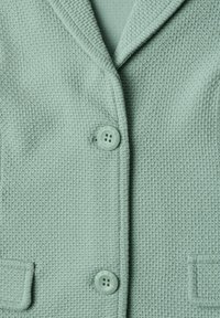 Gros plan sur une veste texturée vert clair avec deux boutons et un col cranté.