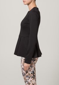 Haut noir à manches longues en tissu texturé, avec un design à basque et un corsage ajusté. Porté avec des leggings à motifs.