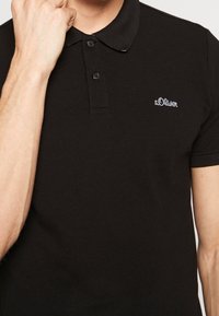Uomo che indossa una polo nera s.Oliver con bottoni, alzando la mano sinistra vicino al volto, ritagliato al collo e con le braccia visibili.