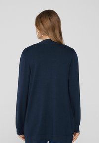 Cárdigan de manga larga azul marino con un corte holgado, textura suave y escote en V. Presenta un dobladillo trasero ligeramente más largo y un detalle mínimo.
