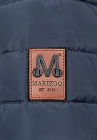 Tumši zila pūkainā jaka ar brūnu ādas etiķeti, kurā ir uzspiests logo "M" un teksts "MARIKOO EST 2016" melnā krāsā. Teksturalizēta virsma.