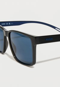 Juoda ir mėlyna stačiakampio formos saulės akiniai su mėlynais objektyvais, turintys matinį paviršių ir iškilų "HUGO" logotipą ant šventyklų.