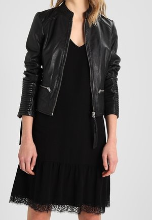 Femme portant une veste en cuir noire avec des détails de fermeture éclair par-dessus une robe noire avec un bord en dentelle à l'ourlet, debout devant un fond uni.
