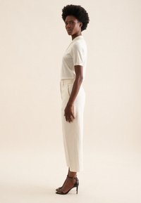 Kremowa bluzka z krótkim rękawem w połączeniu z dopasowanymi białymi spodniami i czarnymi sandałami na obcasie. Gładka faktura i slim fit z elegancką sylwetką.