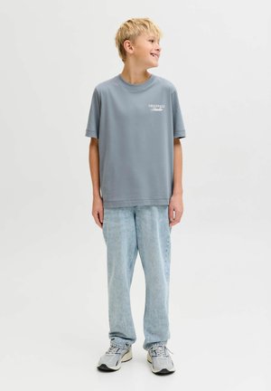 Ragazzo biondo sorridente che guarda verso destra, indossa una t-shirt azzurro chiaro oversize, jeans larghi a lavaggio chiaro e sneaker grigie, su uno sfondo bianco neutro.