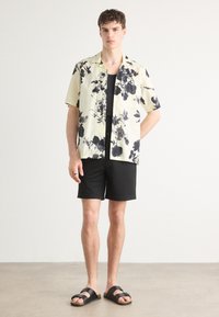 Krátký rukáv, světle žlutá květinová košile s černým monochromatickým potiskem, v kombinaci s černými šortkami a černými sandály. Casual a uvolněný střih.