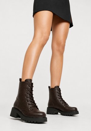 Mujer con botas militares negras de suela gruesa y cordones combinadas con una minifalda negra sobre un fondo claro liso.